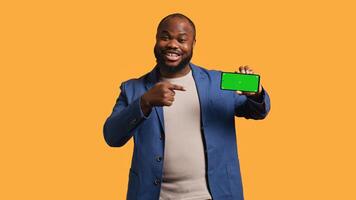 Portrait of man doing influencer marketing using isolated screen phone, showing thumbs up sign, studio background. Merry BIPOC person holding empty copy space mockup cellphone, camera B photo