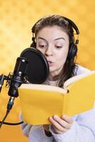 Woman doing voiceover reading of book to produce audiobook using dramatic acting. Narrator using storytelling skills and expressive sounds while producing recording of novel, studio background photo