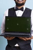 Doorkeeper bellboy holding a laptop with green screen in studio, presenting isolated chroma key display while he works in hospitality industry at a five star hotel. Professional staff. photo