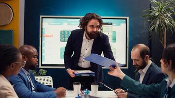 Staff supervisor provides clipboards with current data on performance during an organizational meeting. Lead consults his employees regarding productivity and optimization efforts. Camera B. photo