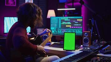 Musical producer learning to tune his guitar before playing, watching lesson tutorial by using greenscreen display on tablet. Young artist tuning strings on instrument, practicing. Camera B. photo