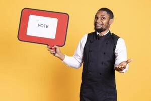 Professional waiter showing a message to vote on speech bubble, creating an advertisement for politics on cardboard icon. Restaurant employee working in customer oriented industry. photo