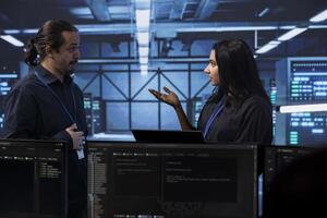 Programmers in server room discussing next steps after checking recovery plan and monitoring energy consumption. Colleagues in data center location talking about routine disk checks results photo