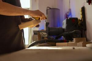 Woodworking professional using manual sandpaper to sander lumber block, making furniture in assembling shop. Carpenter smoothing surface of piece of wood with abrasive material, close up photo