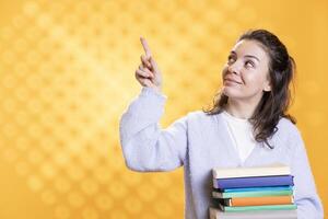 Joyous woman holding pile of books, pointing towards empty copy space, studio background. Cheerful book lover with stack of novels in arms, doing advertisement for literature importance photo
