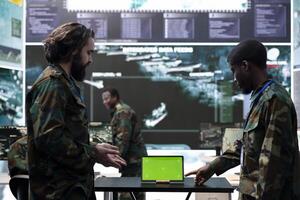 Diverse team of soldiers collecting information from a green screen, employing a tablet in military base of operations. Skilled IT engineers examining activity in control center, high tech. photo