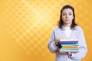 Portrait of woman with worried look on face carrying stack of books, gathering information for school exam, studio background. Student feeling anxious before university assessment, holding textbooks photo