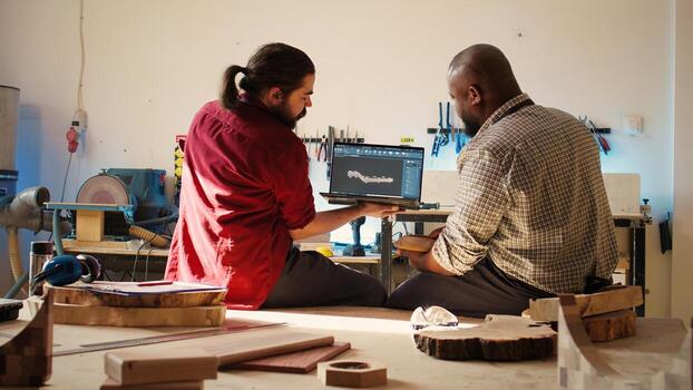 Woodworker looking over blueprints on laptop in CAD software with apprentice. Manufacturer and coworker designing custom 3D furniture on notebook in studio, preparing for customer order, camera B photo