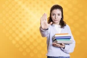 Portrait of woman holding stack of books doing stop sign gesturing, isolated over studio background. Student with pile of textbooks in arms used for academic learning doing halt hand gesture photo