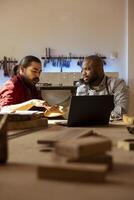 Carpenter and colleague verifying finished wood art product before sending it to client. Artisan and apprentice inspecting wooden object for damages based on laptop checklist photo