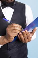 African american hotel porter making a list with room reservations to keep track of bookings and clients, blue background. Receptionist with suit and tie takes notes on papers. Close up. photo