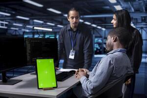 Multiethnic engineering team works on green screen tablet in data center housing storage gear. Diverse staff members using mockup device in server hub with networking components photo