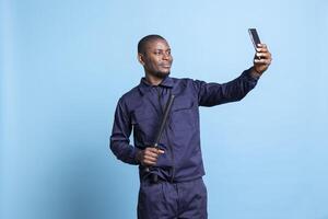Serious security guard enjoys taking pictures with his baton against blue background, posing with weapon on mobile phone camera. Confident agent takes photos in studio on duty.