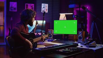 Young artist playing his guitar and practicing new song with online tutorial, looks at computer with greenscreen display. Musician learning new acoustic instrument accords, home studio. Camera B. photo