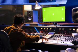 African american audio technician works with mockup layout on pc, adjusting volume levels with knobs on mixing console. Sound designer recording tracks on control desk panel board. photo