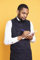 Skilled restaurant server takes the meal order from clients on his notebook, writing the food and drink choice for customer satisfaction. Professional waiter with apron in studio. photo