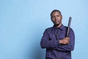 African american security guard feels proud with his baton in studio, posing with confidence with his weapon and professional uniform. Skilled agent standing against blue background. photo