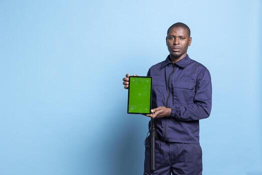 Mall security agent presenting a tablet with isolated mockup screen on camera, wearing work uniform equipped with baton and posing in studio. Bodyguard showing greenscreen display. photo