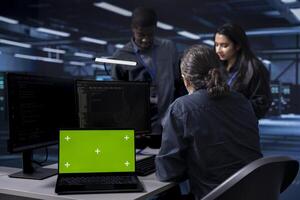 Multiethnic team using green screen screen laptop to check for signs of imminent failure in data center. Diverse staff members working on mockup notebook to prevent damages in server farm photo