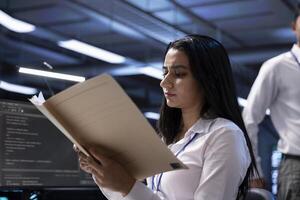 Data center supervisor checking emergency procedures paperwork to ensure company continuity in case of system failure. Employee in server hub facility ensuring compliance with industry regulations photo