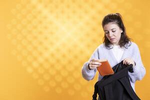 Portrait of woman opening backpack to take encyclopedia book out for school project, isolated over studio background. Student removing textbook from schoolbag, using it for academic purposes photo