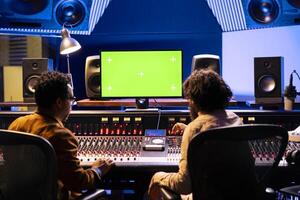 Team of artists mixing and mastering music with greenscreen on pc in professional studio, using audio console with switchers and sliders in control room. Singer and sound technician create songs. photo