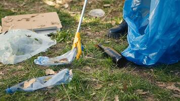 Volunteer using tongs tool to collect trash and plastic waste from the woods, storing rubbish in a garbage bag and clean the natural environment. Activist grabbing litter. Close up. Camera A. photo