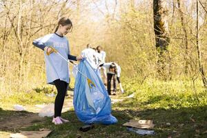 Cheerful proud young kid finishing litter cleanup in a forest, restoring natural environment and helping with forest pollution. Girl recycling plastic waste and using a long claw tool. photo