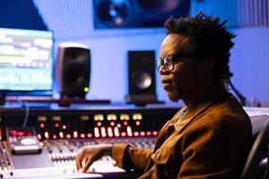 African american record producer working on mixing tunes in music studio, editing sounds in control room with technical equipment and console. Skilled audio expert producing new songs. photo