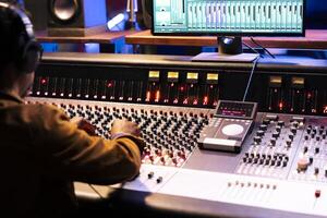 African american sound designer operates control room mixer and audio software for editing new music. Studio technician working with switchers, buttons, and volume level knobs, play hit song. photo