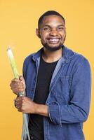 African american person pretending to fight with a leek as a sword, starting combat action with green onion. Smiling man stands against yellow background, playing around with food. photo
