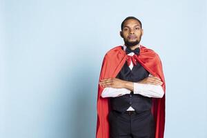 African american doorman posing in a superhero red mantle in studio, acting as a hero and savior for the clients. Happy confident bellhop with a superhuman cape, professionalism. photo