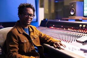 Portrait of african american sound engineer producing records in studio, working with digital audio software and professional mixing console in control room. Expert adjusting volume levels on songs. photo