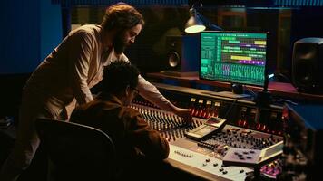 Diverse team of musician and audio technician creating a hit song, working in professional music studio. Artist with producer editing and recording tracks on mixing console panel. Camera B. photo