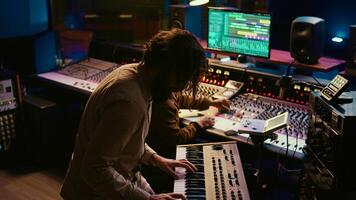 Skilled musician performing in control room at studio and recording tracks for new album, working with audio console and flying faders. Songwriter composing new music on technical tools. Camera B. photo