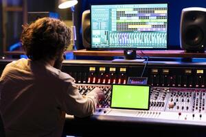 Skilled sound designer recording tunes next to tablet with greenscreen, editing and processing in control room. Music engineer using sliders and buttons to do mix and master., working on an album. photo