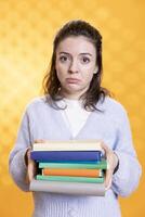 Portrait of woman with distressed facial expression carrying books, afraid of upcoming school exam, studio background. Tense student with textbooks feeling distressed before college assessment photo