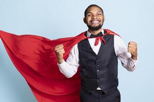 Confident bellboy with suit and tie wearing a red superhero cape, posing as a superhuman and smiling. Young doorkeeper at hotel protecting the guests, hero concept in studio. photo