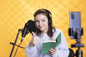 Upbeat media star showing thumbs up while filming vlog at work, reading book to record audiobook. Smiling woman doing content creation, recording novel, studio background, doing positive hand gesture photo