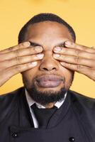 Restaurant worker showing the three wise monkeys gesture in studio, covering his eyes and see no evil as a sign of wisdom and concealment. Butler in apron shows wise symbolic metaphor. photo