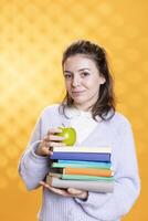 Portrait of radiant woman holding pile of books and organic apple, conveying joy of reading and eating healthy, studio background. Upbeat lady with novels and fresh fruit, harmonious lifestyle concept photo
