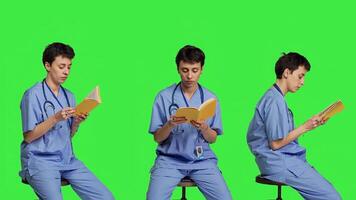 Young nurse studying for the physician exam by reading book in studio, sitting on chair against greenscreen. Specialist enjoys healthcare industry literature, preparing to become a doctor. Camera B. photo