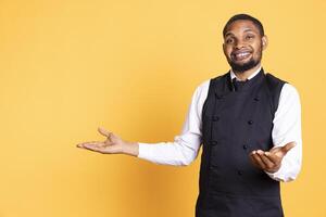 African american waiter points at something to his left or right, indicating direction to the sides in studio. Customer focused employee wears uniform and apron against background. photo