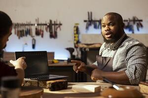 Carpenter in woodworking shop arguing with BIPOC client about commissioned furniture assembling job. Cabinetmaker communicating with customer about blueprint on CAD software on notebook photo