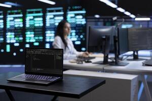 Focus on laptop in front of data center employees in blurry background looking for firewall misconfigurations affecting servers. Close up on notebook used by computer scientists in server room photo