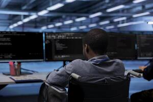African american person with paraplegia working in inclusive server room workplace, typing on PC. Man in wheelchair doing tasks in data center housing advanced data storage infrastructure photo