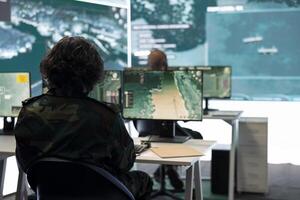 IT operator monitoring surveillance radar footage from CCTV system, using a big screen in military control room. Army recruit collecting information for missions and defense operations. photo