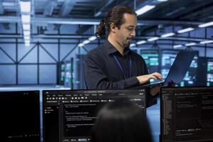 Engineer standing in data center, typing on laptop, doing yearly checkup to avoid equipment downtime. IT employee in server hub monitoring server infrastructure components using tablet photo