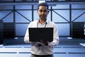 Engineer in server farm checking recovery plan on laptop, monitoring data center energy consumption across components, making sure sensors are functioning optimally, doing maintenance on equipment photo