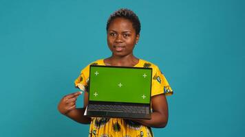 Young adult showing a laptop with isolated mockup screen in studio, presenting a display with green screen against blue background. Female model posing with chroma key. Camera A. photo